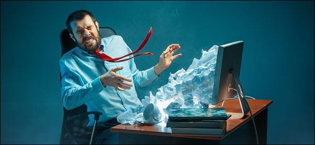 شاب رجل أعمال وسيم مرهق يصرخ على شاشة الكمبيوتر المحمول وغاضبًا من البريد الإلكتروني العشوائي في مكتبه في مكتب حديث. ملصقة بجبال من الورق المجعد.